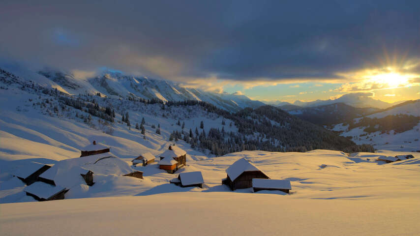 白雪皑皑的山地景观，有几间小屋和日落