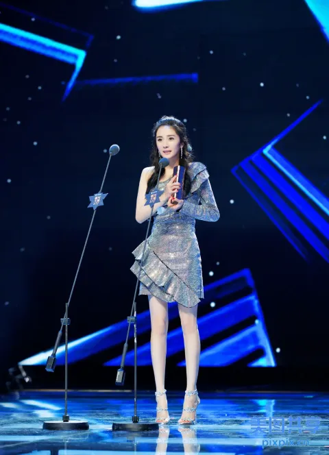 身穿银色连衣裙的阿拉菲女子站在舞台上，拿着麦克风