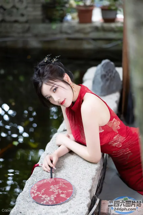 身穿红色连衣裙的 arafed 亚洲女子靠在池塘附近的墙上