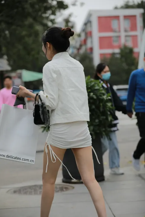 身穿白色夹克和短裤的女子拿着手机走在街上