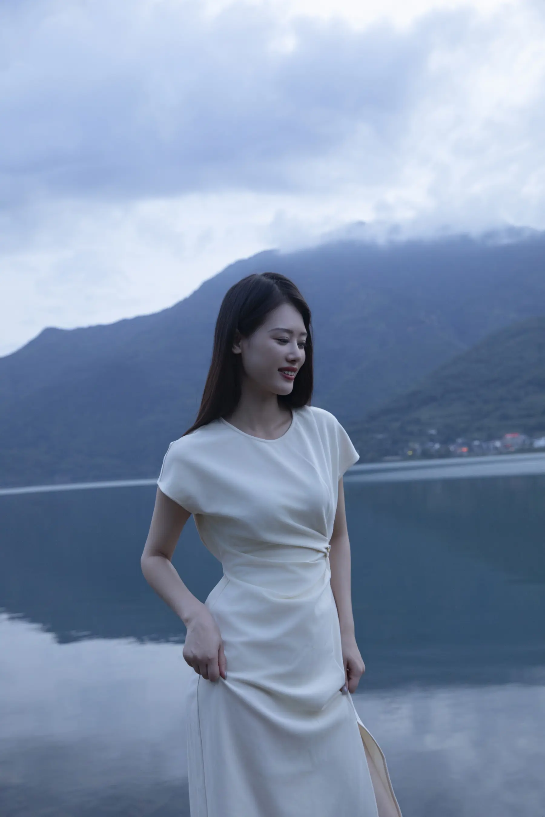 身穿白色连衣裙的 Arafed 女子站在湖边，背景是群山
